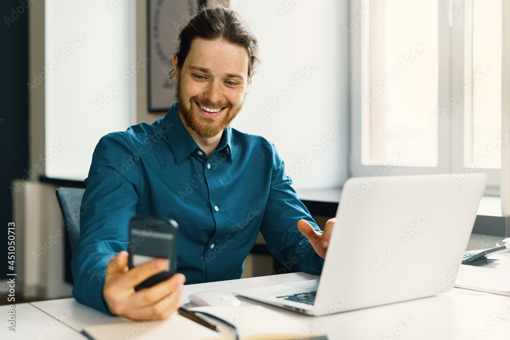 Smiling young guy employee in casual wear looking at smartphone screen, reading good news in message or chatting with friend in social media while working at workplace in modern office Smiling young guy employee in casual wear looking at smartphone screen, reading good news in message or chatting with friend in social media while working at workplace in modern office