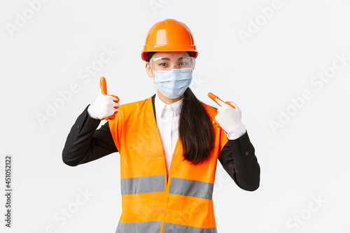 Covid-19 safety protocol at enterpise, construction and preventing virus concept. Asian female chief engineer telling wear face mask at work, pointing at hers and showing thumbs-up in approval photo