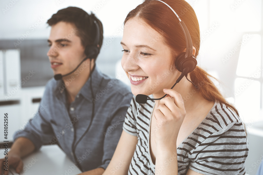 Casual dressed young woman using headset and computer while talking with customers online. Group of operators at work in sunny office. Call center Casual dressed young woman using headset and computer while talking with customers online. Group of operators at work in sunny office. Call center