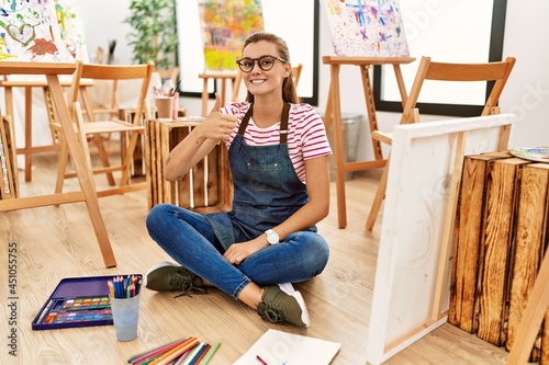 Young brunette woman at art studio sitting on the floor doing happy thumbs up gesture with hand. approving expression looking at the camera showing success.
