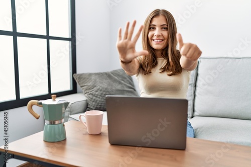 Young brunette woman using laptop at home drinking a cup of coffee showing and pointing up with fingers number six while smiling confident and happy.