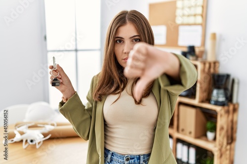 Young brunette woman working at architecture office holding house keys with angry face, negative sign showing dislike with thumbs down, rejection concept