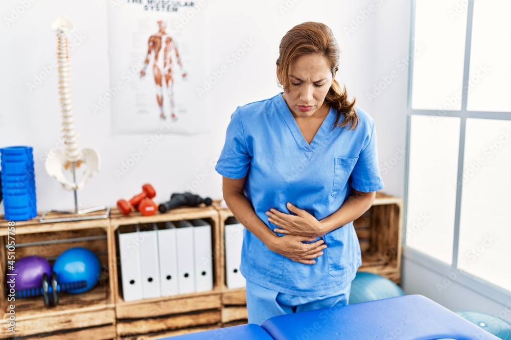 Middle age hispanic physiotherapist woman working at pain recovery clinic with hand on stomach because indigestion, painful illness feeling unwell. ache concept. Middle age hispanic physiotherapist woman working at pain recovery clinic with hand on stomach because indigestion, painful illness feeling unwell. ache concept.