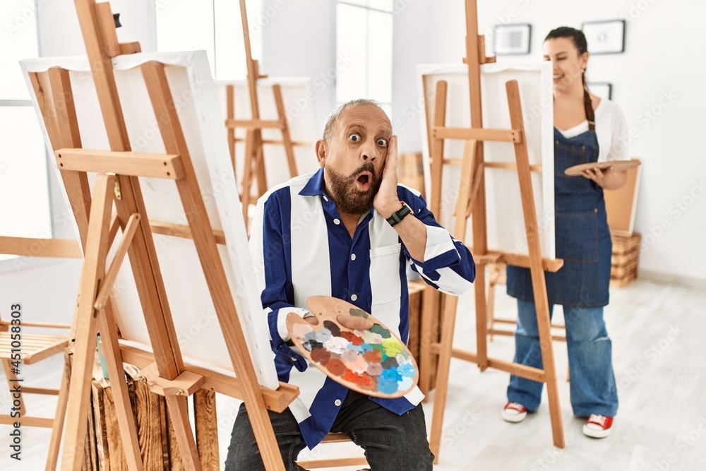 Senior artist man at art studio afraid and shocked, surprise and amazed expression with hands on face Senior artist man at art studio afraid and shocked, surprise and amazed expression with hands on face