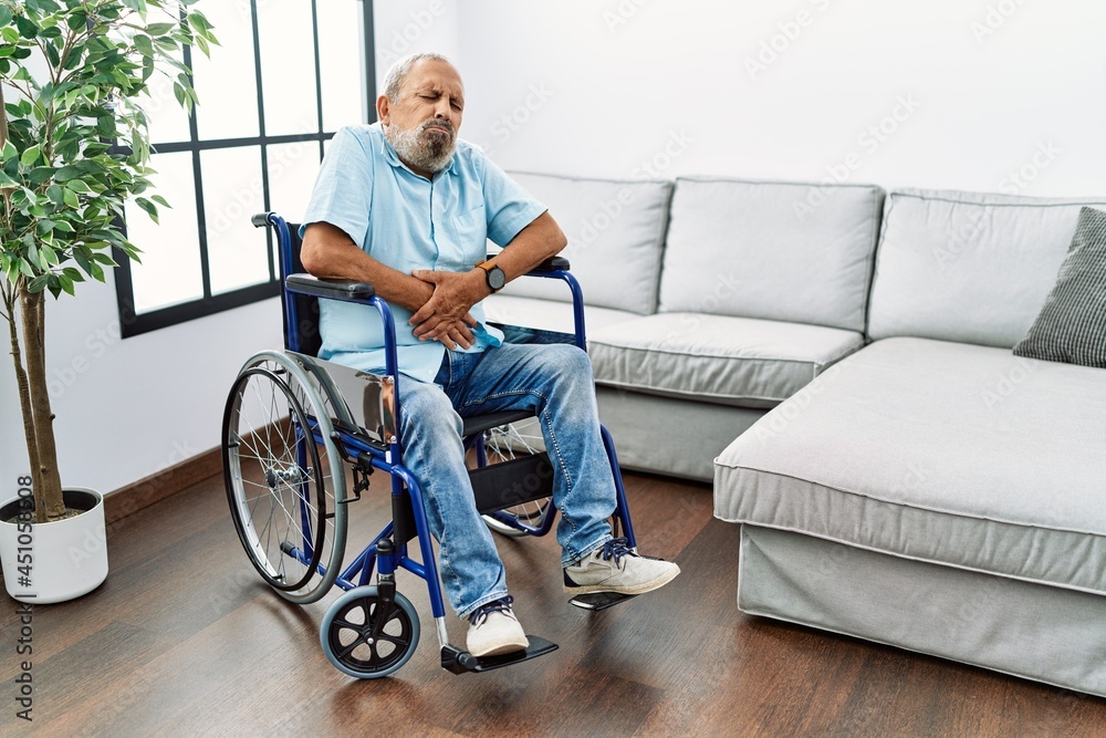 Handsome senior man sitting on wheelchair at the living room with hand on stomach because nausea, painful disease feeling unwell. ache concept. Handsome senior man sitting on wheelchair at the living room with hand on stomach because nausea, painful disease feeling unwell. ache concept.