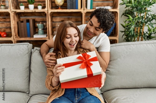 Young couple smiling happy sitting on the sofa surprise with anniversary gift at home.