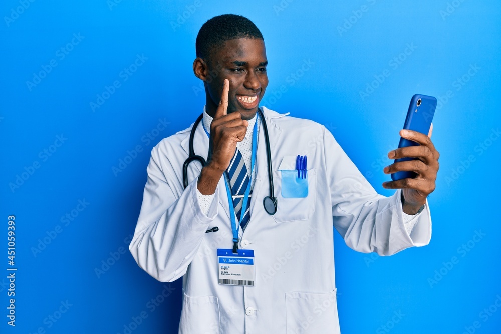 Young african american man wearing doctor uniform using smartphone smiling with an idea or question pointing finger with happy face, number one Young african american man wearing doctor uniform using smartphone smiling with an idea or question pointing finger with happy face, number one