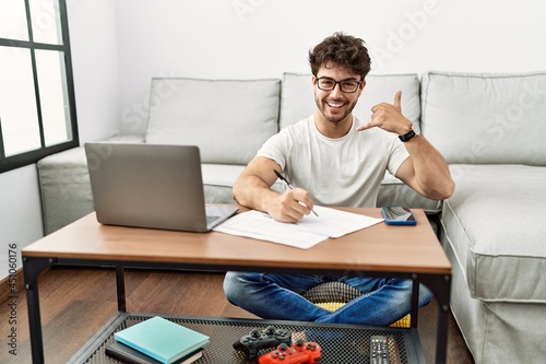 Hispanic man doing papers at home smiling doing phone gesture with hand and fingers like talking on the telephone. communicating concepts.