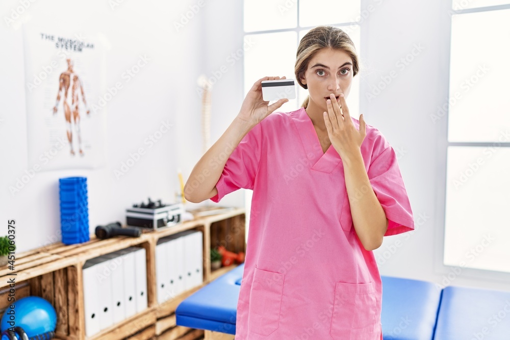 Young physiotherapist woman working at pain recovery clinic holding credit card covering mouth with hand, shocked and afraid for mistake. surprised expression