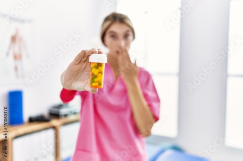 Young physiotherapist woman working at pain recovery clinic holding pills covering mouth with hand  shocked and afraid for mistake. surprised expression