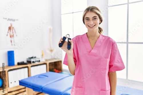 Young blonde woman working at pain recovery clinic holding hand strengthener looking positive and happy standing and smiling with a confident smile showing teeth
