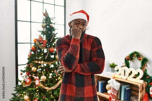 African american man wearing santa claus hat standing by christmas tree looking stressed and nervous with hands on mouth biting nails. anxiety problem.