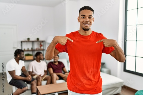 Young african man at home with friends sitting on the sofa at home looking confident with smile on face  pointing oneself with fingers proud and happy.