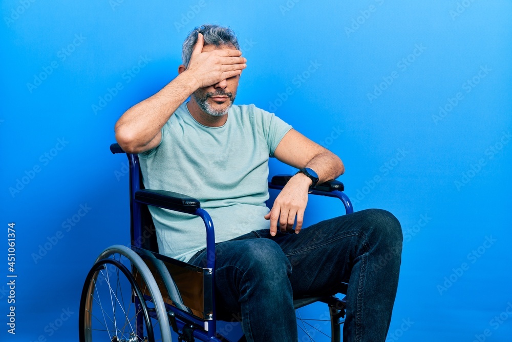 Fototapeta premium Handsome middle age man with grey hair sitting on wheelchair covering eyes with hand, looking serious and sad. sightless, hiding and rejection concept