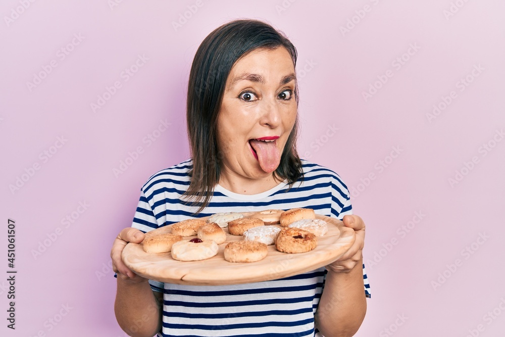Middle age hispanic woman holding sweet pastries sticking tongue out happy with funny expression. Middle age hispanic woman holding sweet pastries sticking tongue out happy with funny expression.