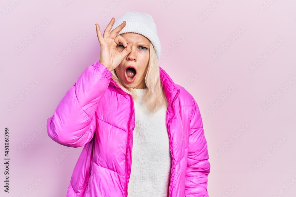 Beautiful caucasian blonde woman wearing wool hat and winter coat doing ok gesture shocked with surprised face, eye looking through fingers. unbelieving expression. Beautiful caucasian blonde woman wearing wool hat and winter coat doing ok gesture shocked with surprised face, eye looking through fingers. unbelieving expression.