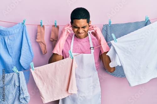 Young handsome hispanic man wearing cleaner apron holding clothes on clothesline covering ears with fingers with annoyed expression for the noise of loud music. deaf concept.
