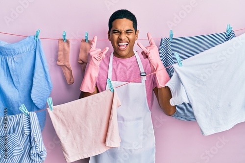 Young handsome hispanic man wearing cleaner apron holding clothes on clothesline shouting with crazy expression doing rock symbol with hands up. music star. heavy concept.