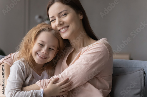 Smiling young Caucasian mother and small teen daughter rest on sofa at home, hug and cuddle enjoy family weekend together. Happy mom and little girl child embrace show love and care in relations.