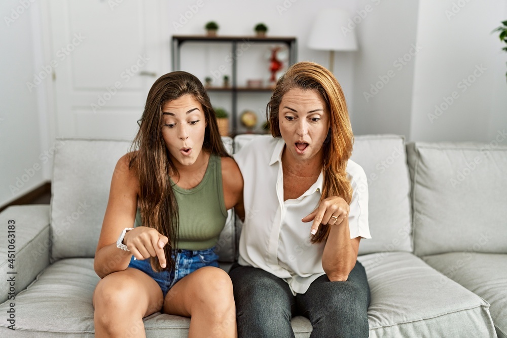 Mother and daughter together sitting on the sofa at home pointing down with fingers showing advertisement, surprised face and open mouth Mother and daughter together sitting on the sofa at home pointing down with fingers showing advertisement, surprised face and open mouth