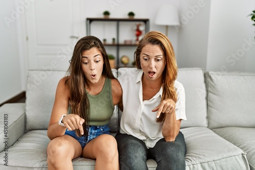 Mother and daughter together sitting on the sofa at home pointing down with fingers showing advertisement, surprised face and open mouth