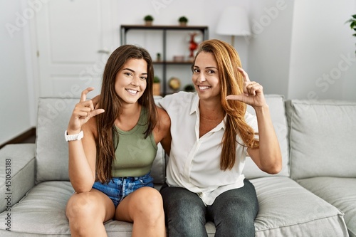 Mother and daughter together sitting on the sofa at home smiling and confident gesturing with hand doing small size sign with fingers looking and the camera. measure concept.