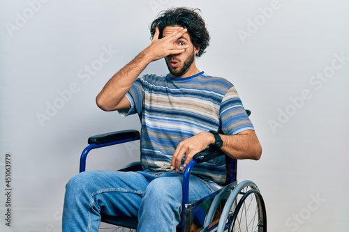 Handsome hispanic man sitting on wheelchair peeking in shock covering face and eyes with hand, looking through fingers with embarrassed expression.