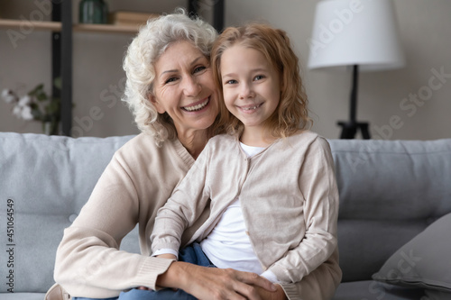 Portrait of happy old Caucasian 60s grandmother and small teen granddaughter sit rest on sofa at home together. Smiling mature grandma and little grandchild cuddle show love and care in relations.