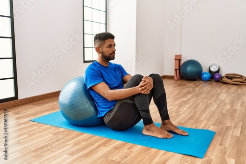 Handsome hispanic man doing exercise and stretching on yoga mat, practicing flexibility with pilates ball at the gym
