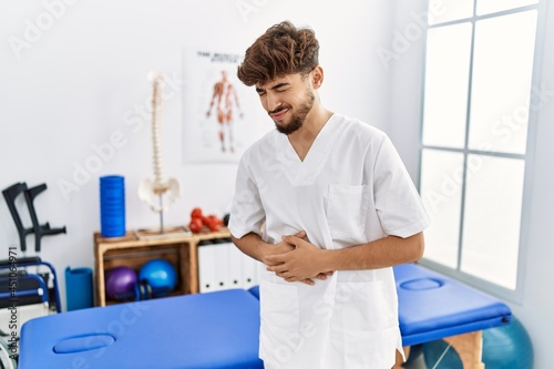 Young arab man working at pain recovery clinic with hand on stomach because nausea  painful disease feeling unwell. ache concept.