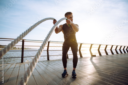 Young man wearing sports clothes is doing exercisesat the beach pier in the morning. Battle ropes. Kettlebell. Sport, Active life.