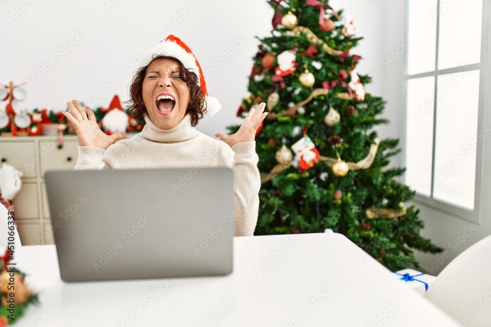 Middle age hispanic woman using laptop sitting by christmas tree celebrating mad and crazy for success with arms raised and closed eyes screaming excited. winner concept Middle age hispanic woman using laptop sitting by christmas tree celebrating mad and crazy for success with arms raised and closed eyes screaming excited. winner concept