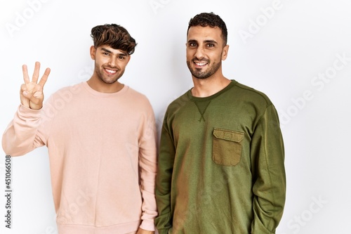 Young gay couple standing over isolated white background showing and pointing up with fingers number three while smiling confident and happy.