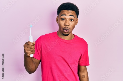 Young african american man holding electric toothbrush scared and amazed with open mouth for surprise, disbelief face