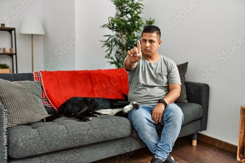 Young latin man and dog sitting on the sofa at home pointing with finger up and angry expression, showing no gesture © Krakenimages.com
