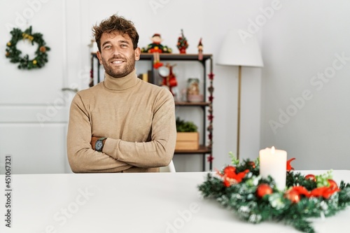 Young handsome man with beard sitting on the table by christmas decoration happy face smiling with crossed arms looking at the camera. positive person. © Krakenimages.com