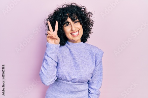 Young middle east woman wearing casual clothes smiling with happy face winking at the camera doing victory sign. number two.