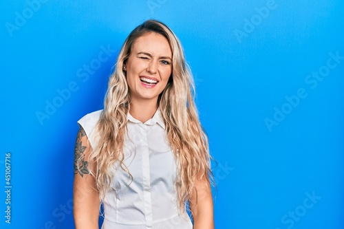 Beautiful young blonde woman wearing casual white shirt winking looking at the camera with sexy expression, cheerful and happy face.