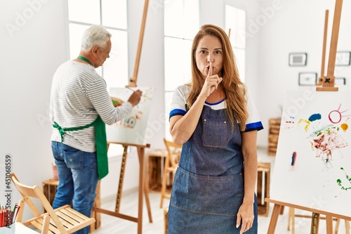 Hispanic woman wearing apron at art studio asking to be quiet with finger on lips. silence and secret concept.
