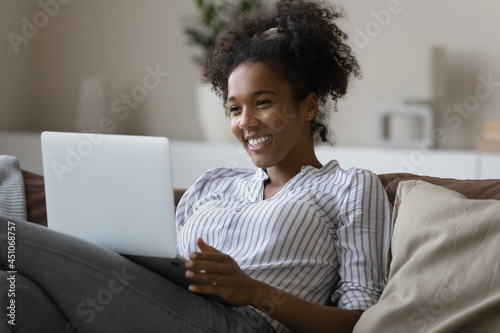 Smiling African American woman relax on sofa talk speak on video call on laptop. Happy young ethnic female have fun laugh engaged in webcam digital virtual event on computer. Technology concept. photo