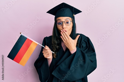 Young hispanic woman wearing graduation uniform holding germany flag covering mouth with hand, shocked and afraid for mistake. surprised expression
