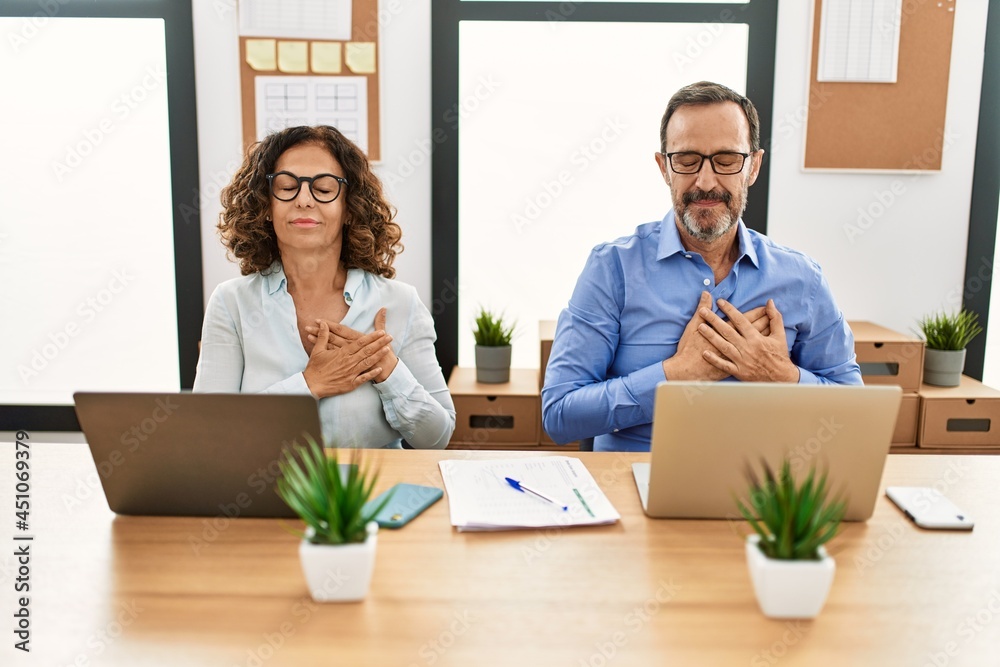 Middle age hispanic woman and man sitting with laptop at the office smiling with hands on chest with closed eyes and grateful gesture on face. health concept. Middle age hispanic woman and man sitting with laptop at the office smiling with hands on chest with closed eyes and grateful gesture on face. health concept.