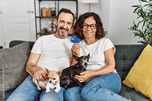Middle age hispanic couple smiling happy and holding credit card. Sitting on the sofa with dogs at home. © Krakenimages.com