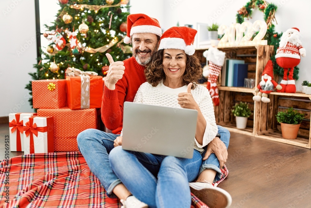Hispanic middle age woman and mature man using laptop sitting by christmas tree smiling happy and positive, thumb up doing excellent and approval sign Hispanic middle age woman and mature man using laptop sitting by christmas tree smiling happy and positive, thumb up doing excellent and approval sign