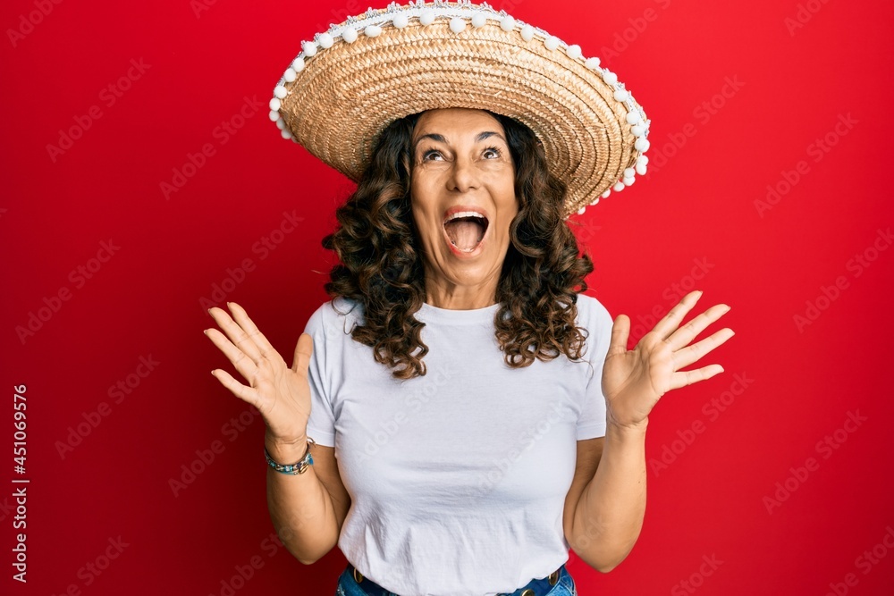 Middle age hispanic woman holding mexican hat celebrating mad and crazy for success with arms raised and closed eyes screaming excited. winner concept