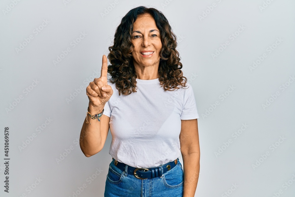 Middle age hispanic woman wearing casual white t shirt showing and pointing up with finger number one while smiling confident and happy. Middle age hispanic woman wearing casual white t shirt showing and pointing up with finger number one while smiling confident and happy.