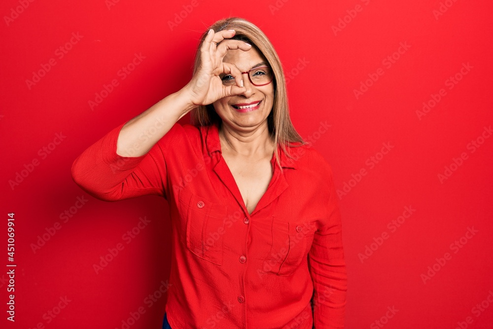 Middle age hispanic woman wearing casual clothes and glasses smiling happy doing ok sign with hand on eye looking through fingers
