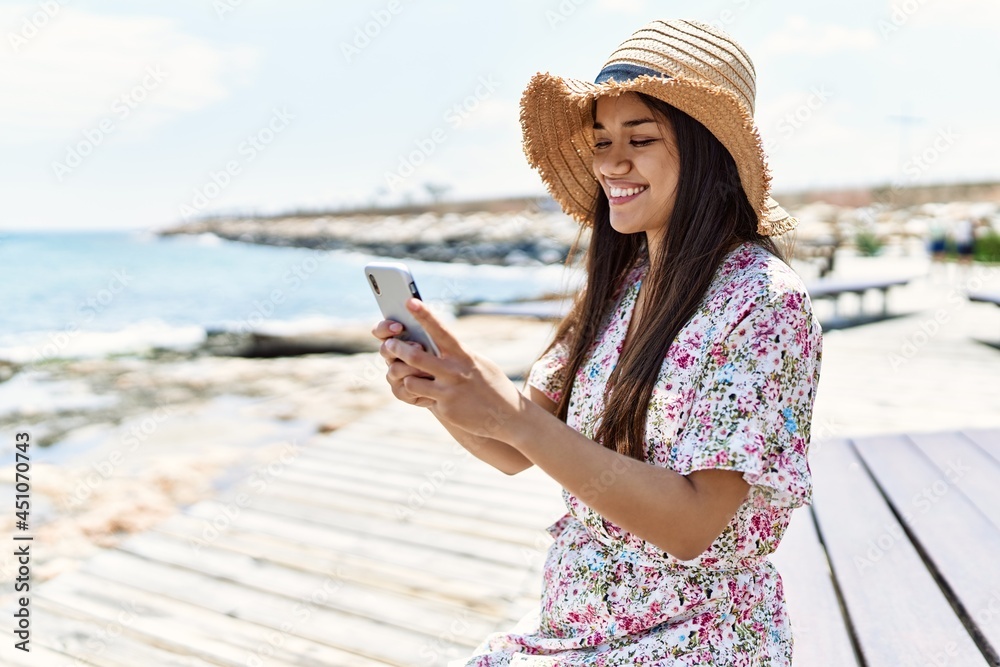 Young latin girl wearing summer using smartphone sitting on the bench hat at the beach.