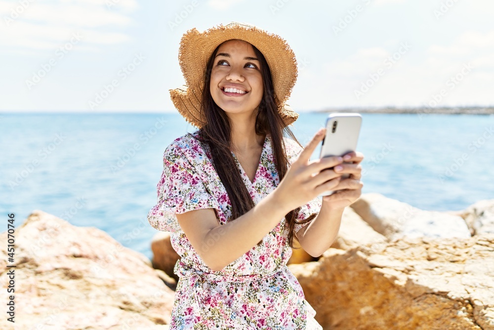 Fototapeta premium Young latin girl wearing summer hat using smartphone at the beach.