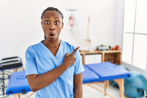 Black woman with braids working at pain recovery clinic surprised pointing with finger to the side, open mouth amazed expression.